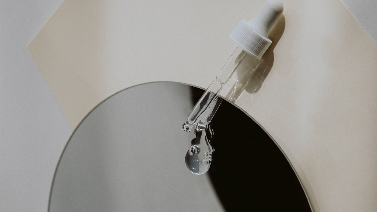 Minimalist shot of a clear glass serum dropper releasing product over a circular mirror, captured in soft neutral tones with natural light reflections.