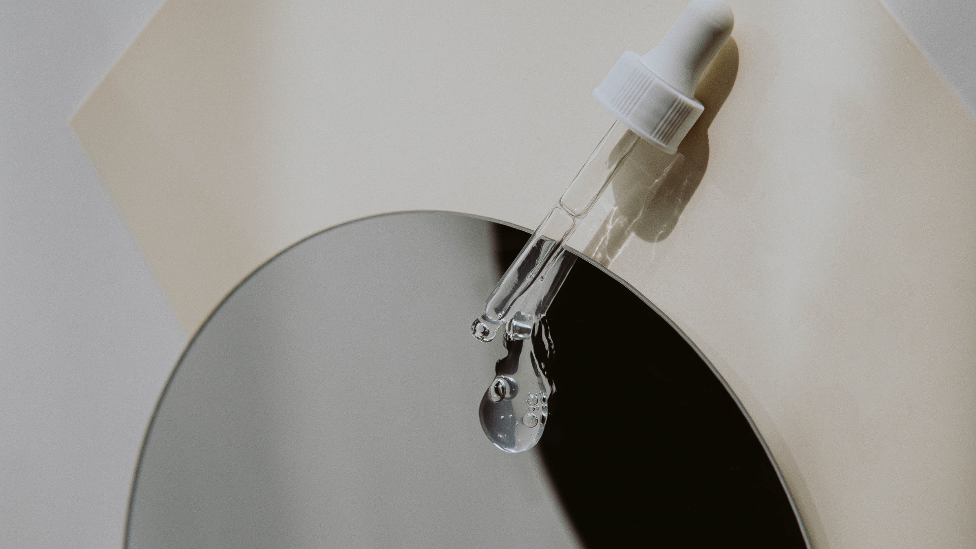 Minimalist shot of a clear glass serum dropper releasing product over a circular mirror, captured in soft neutral tones with natural light reflections.