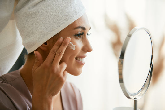 A woman with a towel wrapped around her head is smiling as she applies a moisturizing cream to her cheek. She is looking into a round mirror, enjoying her skincare routine. The setting is bright and serene, suggesting a fresh and healthy start to her day.