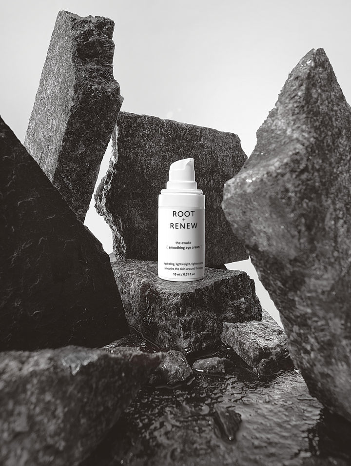 The Awake Smoothing Eye Cream by Root + Renew placed between rugged dark stones and water, evoking strength and resilience.