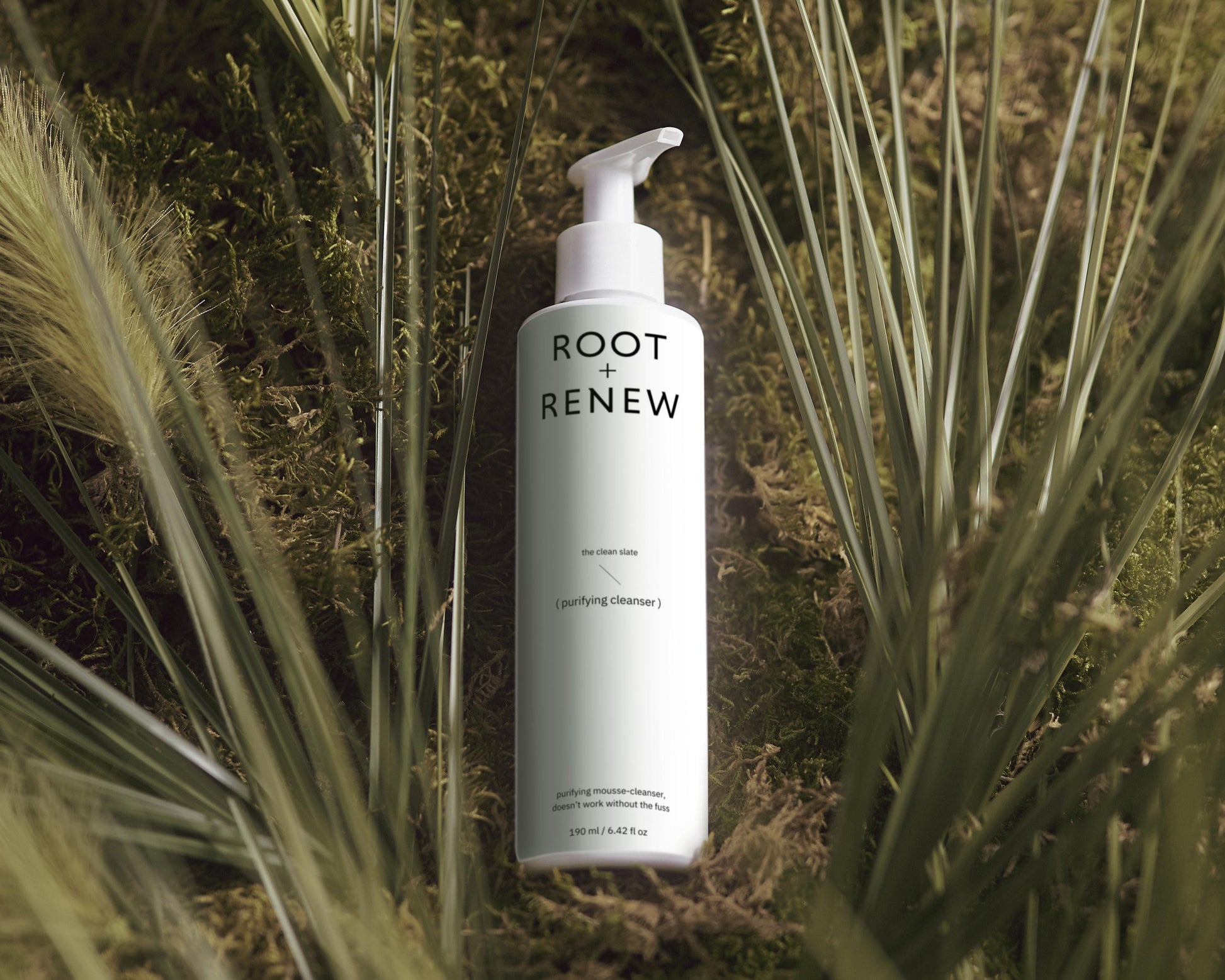 A white bottle of Root + Renew Purifying Cleanser with a pump dispenser stands upright among lush green moss and tall grass-like plants. The minimalistic packaging features black typography and a small blue dot. The label reads "The Clean Slate" and "Purifying Cleanser," indicating a mousse-based facial cleanser. The bottle is positioned as the focal point, surrounded by an earthy, nature-inspired background, emphasizing a clean and natural skincare approach.