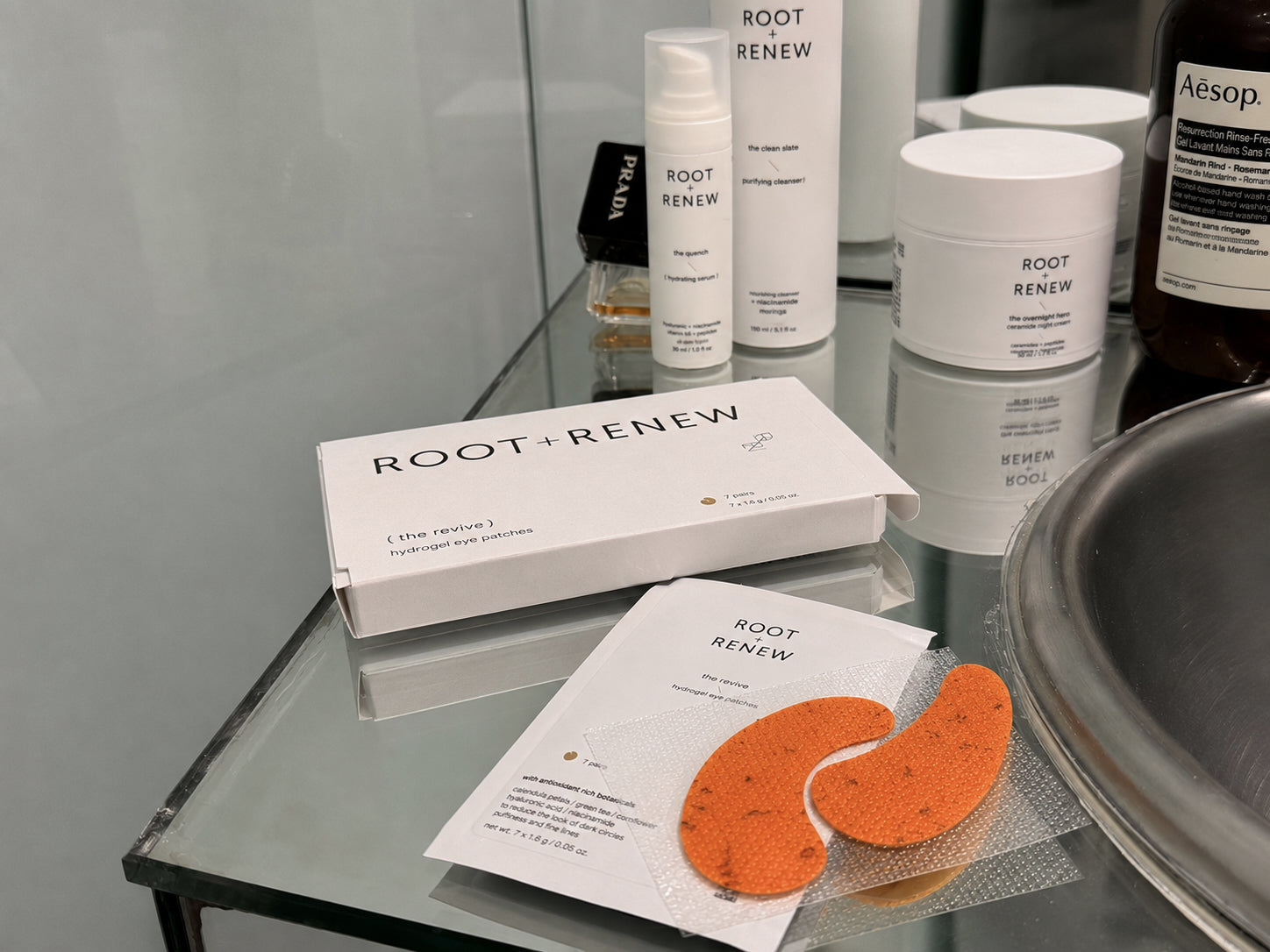 Root + Renew The Revive Hydrogel Eye Patches  skincare products arranged on a glass shelf, including a white product box, a single-use packet with orange eye patches, and several white bottles and jars. An Aesop bottle is partially visible in the background.