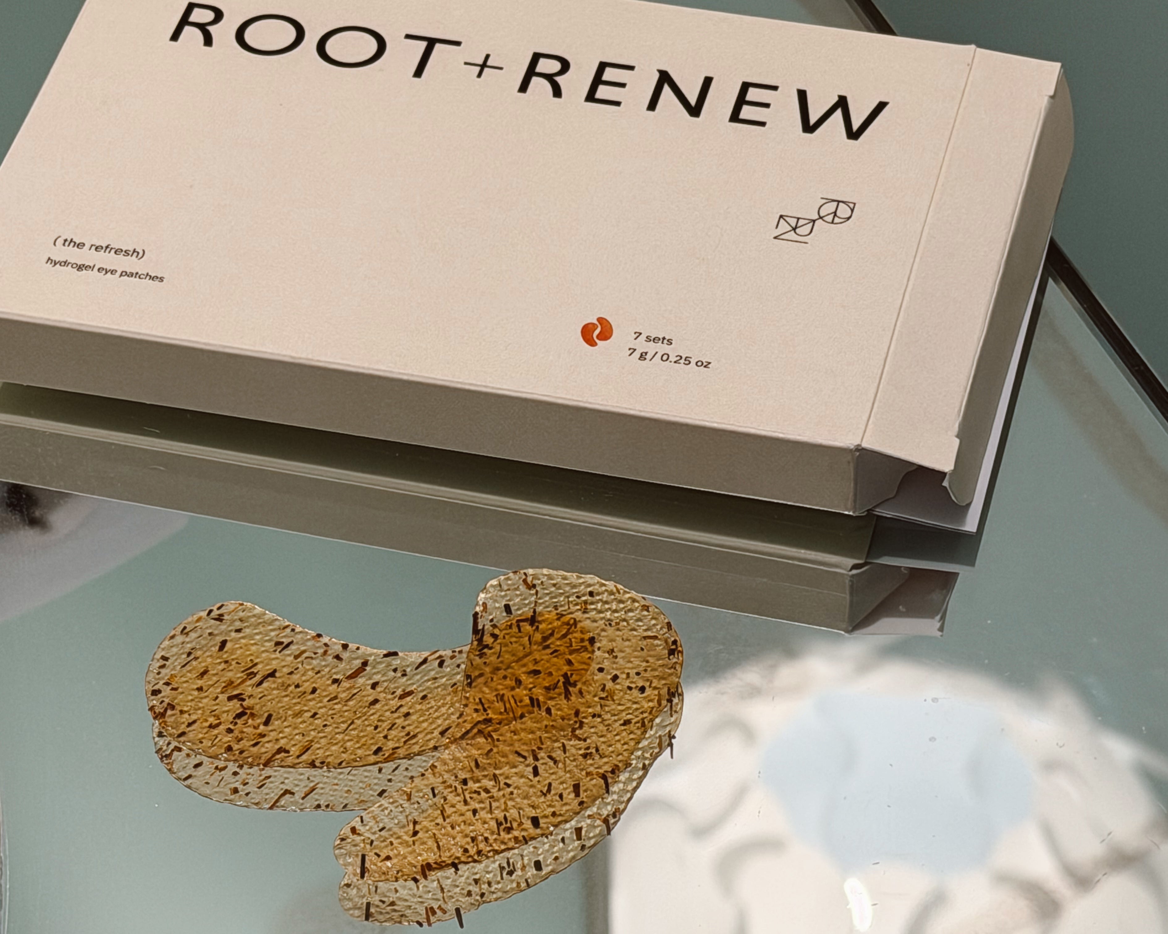 A boxed skincare product labeled ROOT+RENEW sits on a reflective glass surface, with a small honeycomb-style beige and.